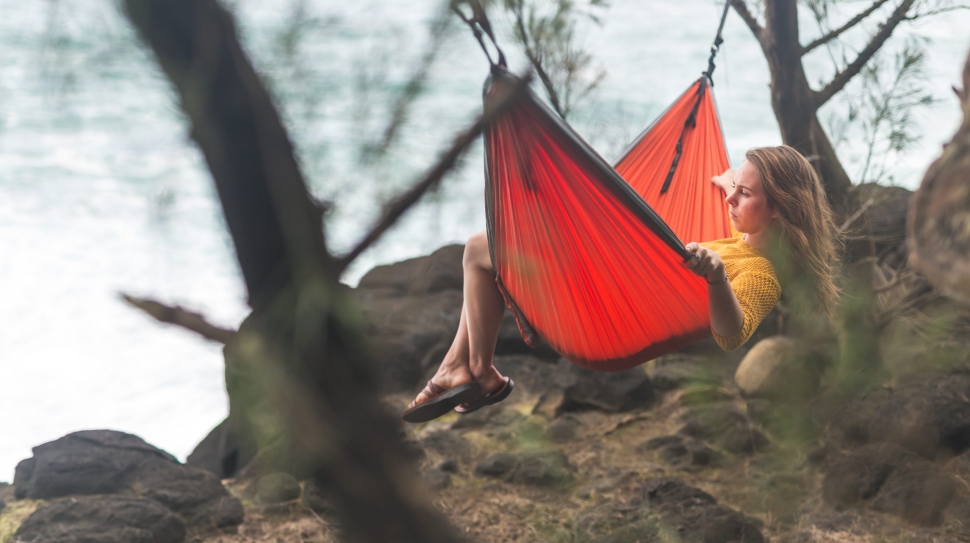 A young Caucasian female relaxes in a hammock suspended between two trees that is overlooking the Pacific Ocean. She is on vacation and enjoying the solitude and rocky coastline of Hawaii as she gazes out at the water.